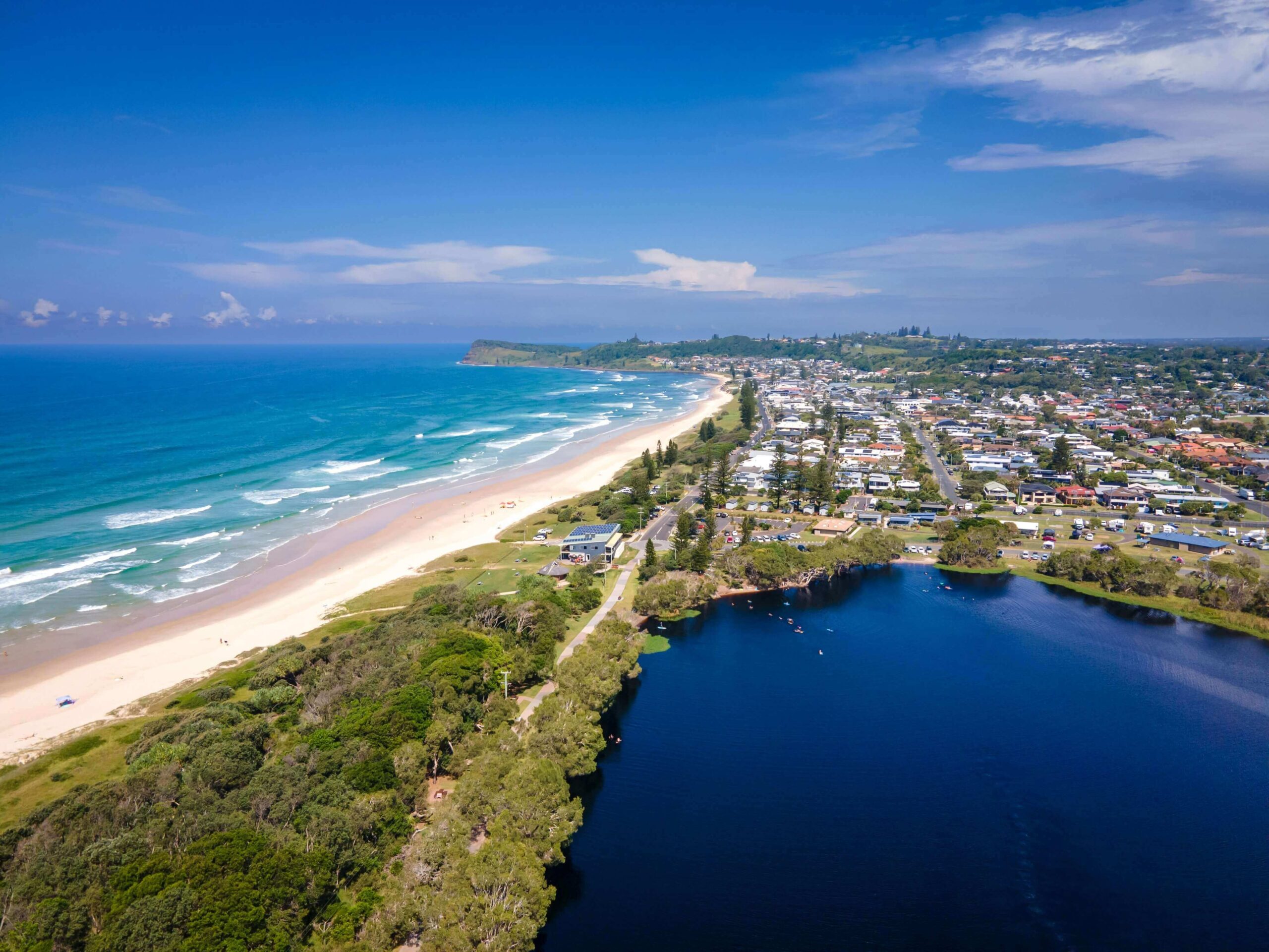 Aerial view of Lennox Head, NSW, showing the dark tea-tree waters of Lake Ainsworth sitting directly beside the white sand and ocean waves of Seven Mile Beach.