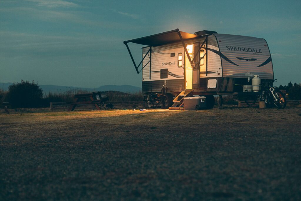 A caravan parked at a campsite at dusk with its side awning extended, illuminated by warm light coming from the open door.
