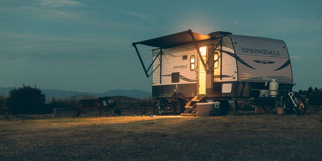 A caravan parked at a campsite at dusk with its side awning extended, illuminated by warm light coming from the open door.