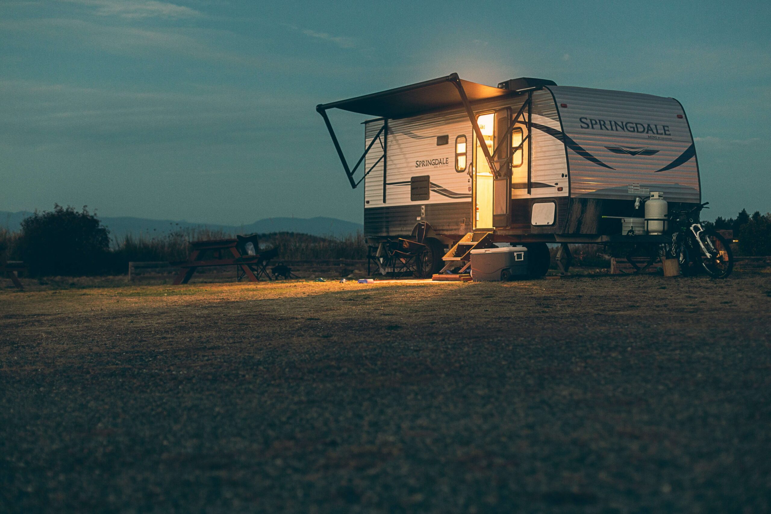 A caravan parked at a campsite at dusk with its side awning extended, illuminated by warm light coming from the open door.